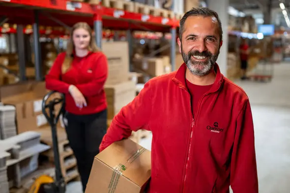 Un homme souriant portant un colis se tient au premier plan, vêtu d'un sweat rouge. À l'arrière, une femme en rouge regarde vers l'objectif, près d'un chariot élévateur. L'environnement est un entrepôt avec des boîtes empilées.