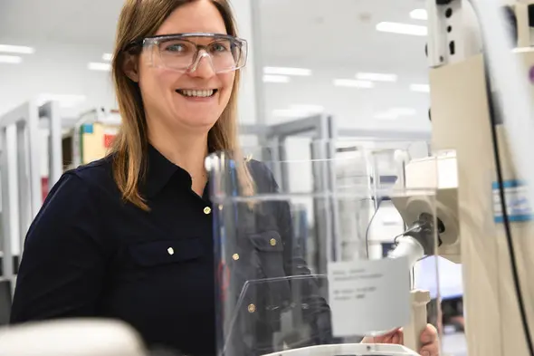 Femme souriante portant des lunettes de protection, se tenant près d'un équipement de laboratoire. Elle est habillée d'une chemise noire et se trouve dans un environnement de travail scientifique.
