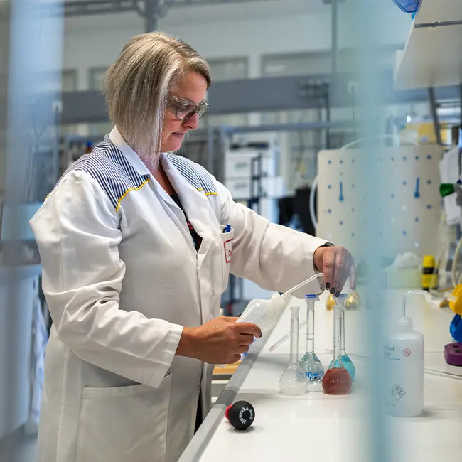 Une scientifique en blouse de laboratoire manipule des instruments et des flacons dans un laboratoire. Elle semble concentrée sur son travail, entourée d'équipements de laboratoire et de divers matériaux scientifiques.