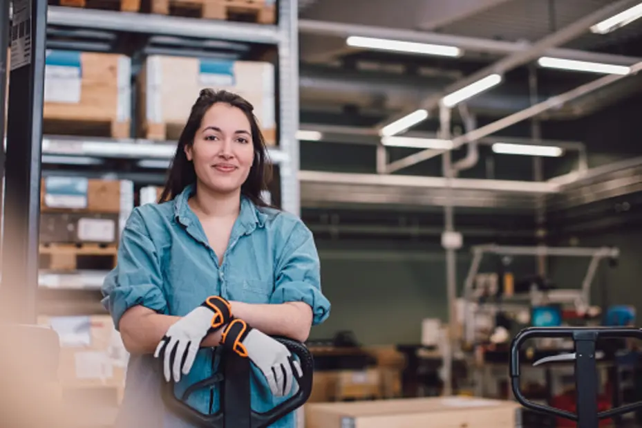 Femme souriante portant des gants de travail, debout près d'un transpalette dans un environnement de stockage. En arrière-plan, des palettes en bois et divers équipements industrielles sont visibles.