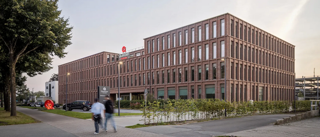 Un bâtiment moderne en briques avec de nombreuses fenêtres, entouré d'espaces verts et de voitures. Des piétons marchent sur le trottoir à l'avant. Un panneau indiquant une entreprise est visible à l'entrée.
