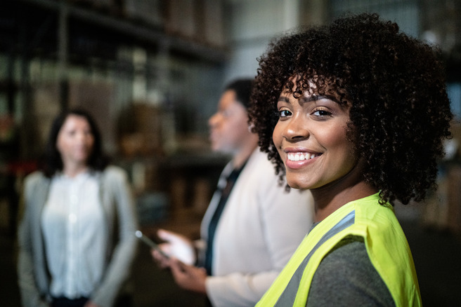 Une femme souriante avec des cheveux bouclés porte un gilet de sécurité dans un entrepôt. Deux autres femmes, en arrière-plan, discutent. L'environnement est industriel, avec des étagères remplies de marchandises.