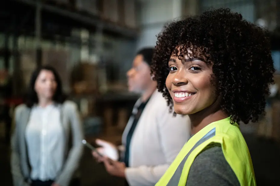 Une femme souriante avec des cheveux bouclés porte un gilet de sécurité dans un entrepôt. Deux autres femmes, en arrière-plan, discutent. L'environnement est industriel, avec des étagères remplies de marchandises.