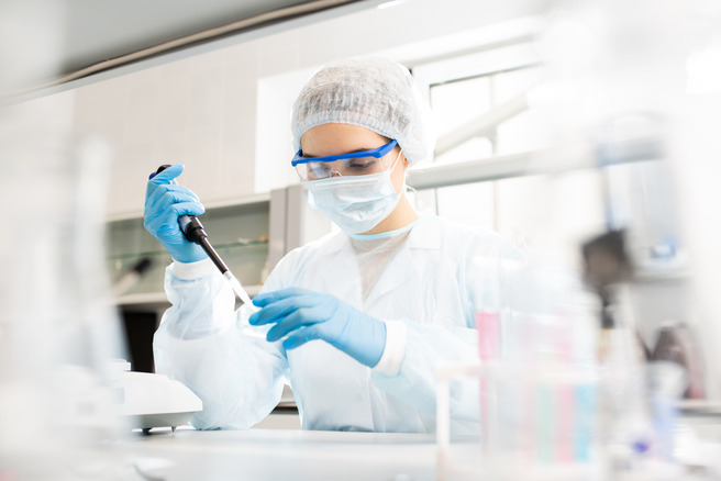Une scientifique en blouse blanche, portant un masque, des lunettes et des gants, utilise un micropipette dans un laboratoire. Elle se concentre sur son travail, entourée de matériel de laboratoire.