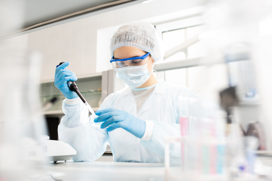 Une scientifique en blouse blanche, portant un masque, des lunettes et des gants, utilise un micropipette dans un laboratoire. Elle se concentre sur son travail, entourée de matériel de laboratoire.