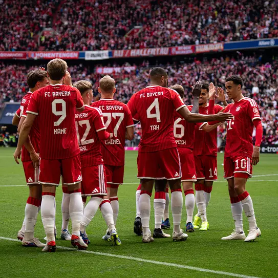 Des joueurs de l'équipe de football du Bayern Munich se regroupent sur le terrain, échangeant des gestes amicaux et des encouragements. Ils portent des maillots rouges avec le logo du club, et un grand nombre de spectateurs peut être vu en arrière-plan.
