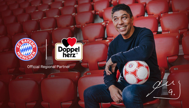 Un homme souriant est assis dans un stade de football, tenant un ballon. Des logos du FC Bayern München et de Doppelherz sont visibles, avec une mention « Partenaire régional officiel ». L'arrière-plan montre des sièges rouges vides.