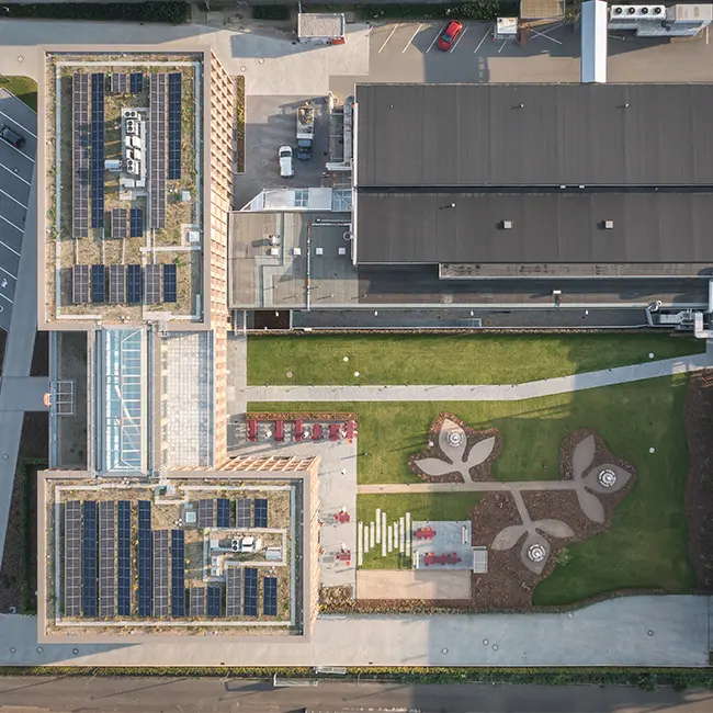 Vue aérienne d'un complexe architectural moderne avec des toits équipés de panneaux solaires. En bas, un aménagement paysager avec des allées et des espaces verts, incluant un jardin stylisé. Des voitures sont stationnées à proximité.