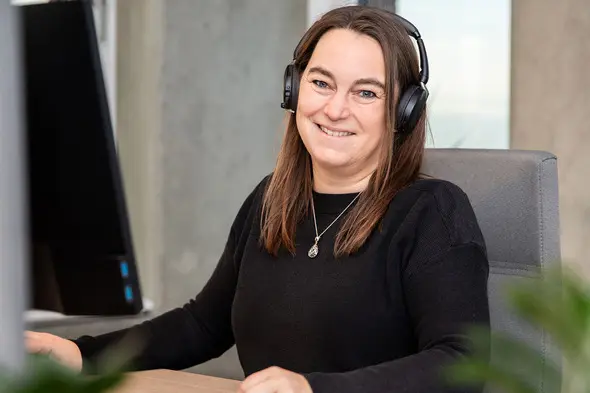 Femme souriante assise à un bureau, portant un casque audio. Elle est face à un ordinateur et a les cheveux longs et bruns. L'environnement de travail est moderne et lumineux, avec des éléments de plantes vertes en arrière-plan.