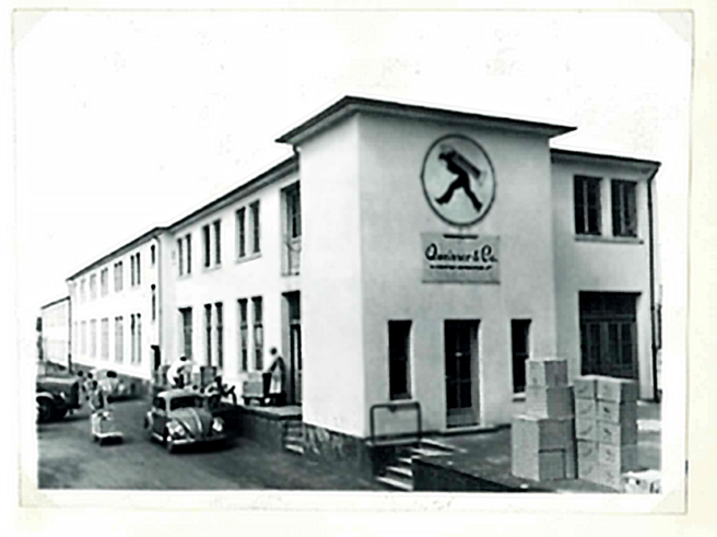 Bâtiment d'une entreprise avec un grand logo représentant une silhouette en mouvement. Des personnes et des voitures anciennes sont visibles à l'extérieur. Des caisses sont empilées sur le sol devant l'entrée. Photo en noir et blanc.