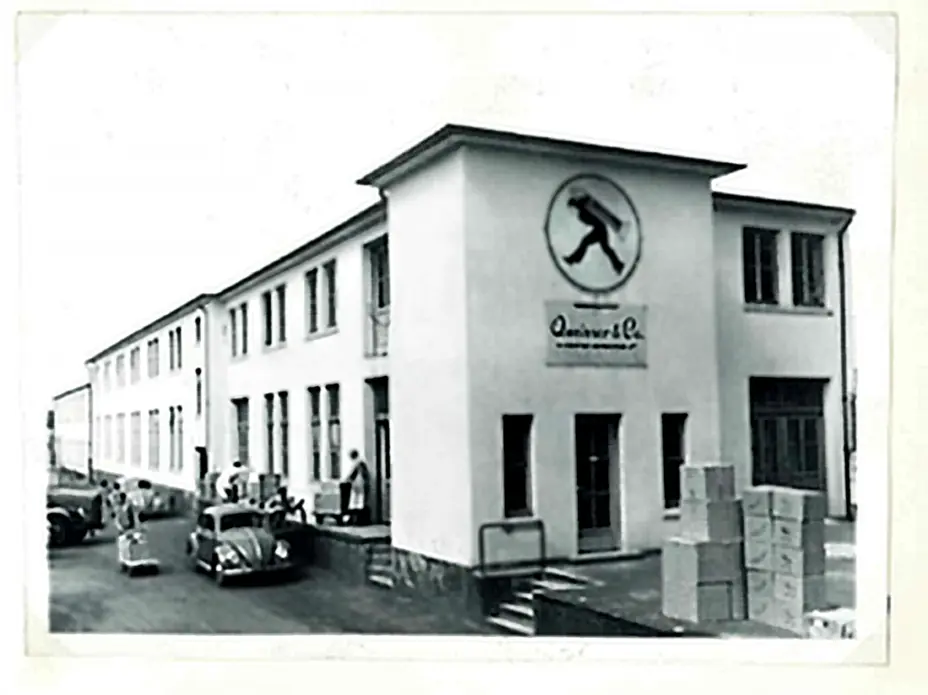 Bâtiment d'une entreprise avec un grand logo représentant une silhouette en mouvement. Des personnes et des voitures anciennes sont visibles à l'extérieur. Des caisses sont empilées sur le sol devant l'entrée. Photo en noir et blanc.