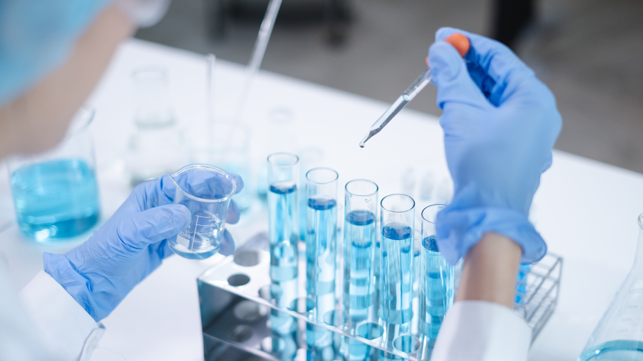 Un scientifique en blouse de laboratoire et gants bleus utilise une pipette pour ajouter un liquide bleu dans des tubes à essai. Il tient également un erlenmeyer contenant un liquide similaire. L'environnement semble être un laboratoire.