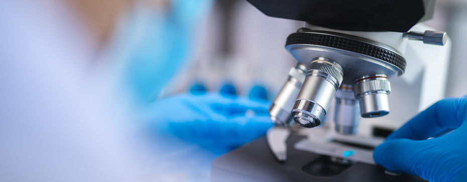 Un gros plan sur un microscope de laboratoire, avec des mains portant des gants bleus manipulant un échantillon. L'arrière-plan flou suggère un environnement de recherche scientifique.