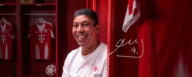 Un homme souriant est assis dans un vestiaire aux nuances de rouge, avec des maillots de football affichés en arrière-plan. Un ballon de football est visible près de lui. Une signature est inscrite en bas à droite.