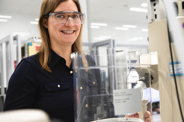 Femme souriante portant des lunettes de protection, se tenant près d'un équipement de laboratoire. Elle est habillée d'une chemise noire et se trouve dans un environnement de travail scientifique.